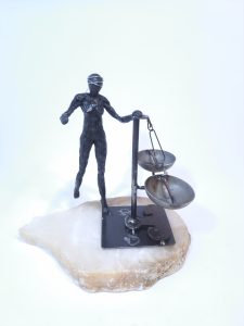 Escultura formada por una figura femenina de resina epoxi con los ojos vendados que sujeta una balanza en equilibro sobre una base de alabastro blanco.