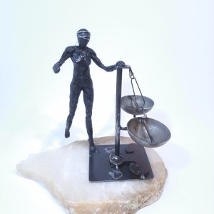 Escultura formada por una figura femenina de resina epoxi con los ojos vendados que sujeta una balanza en equilibro sobre una base de alabastro blanco.