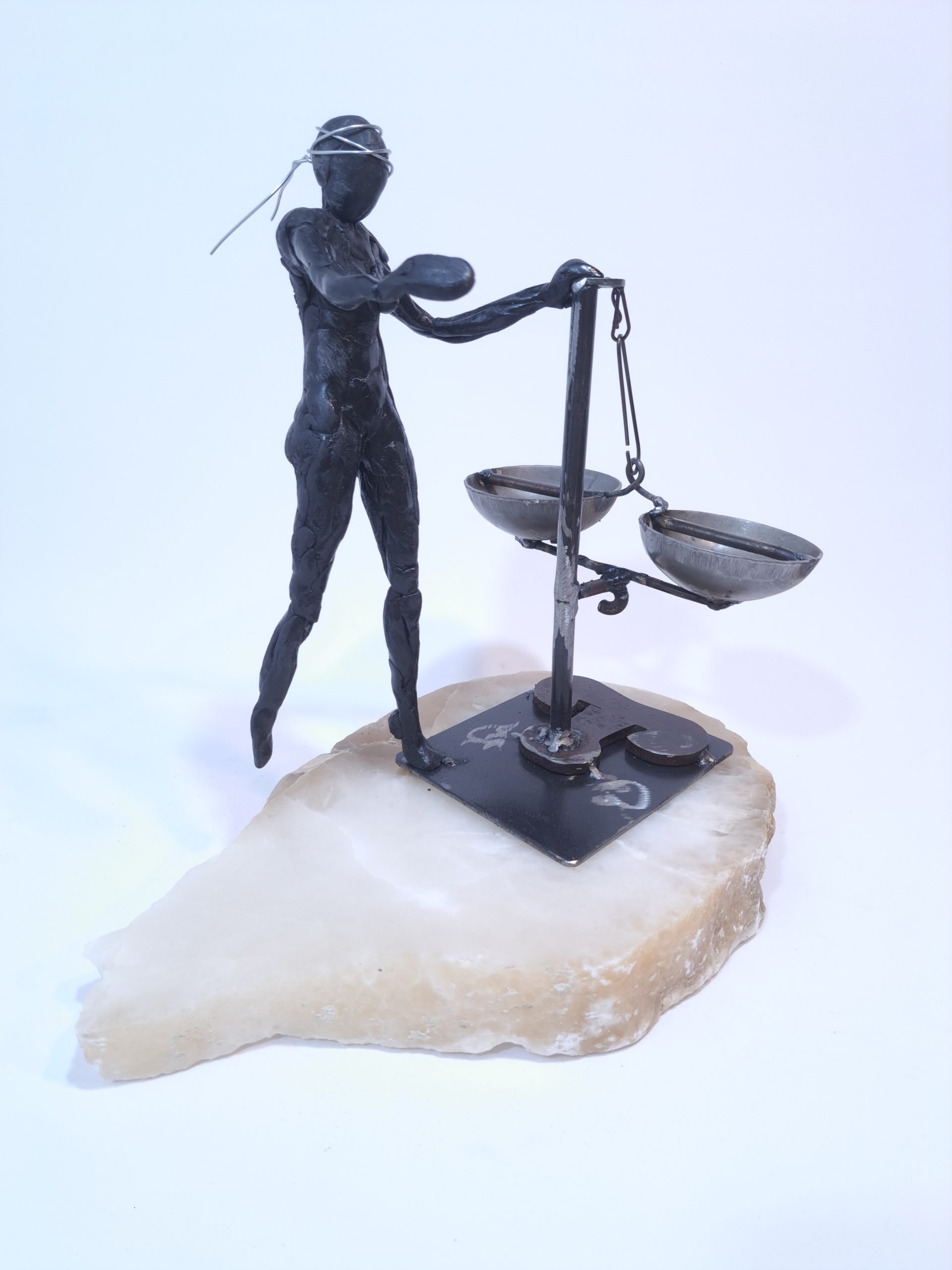 Escultura formada por una figura femenina de resina epoxi con los ojos vendados que sujeta una balanza en equilibro sobre una base de alabastro blanco.
