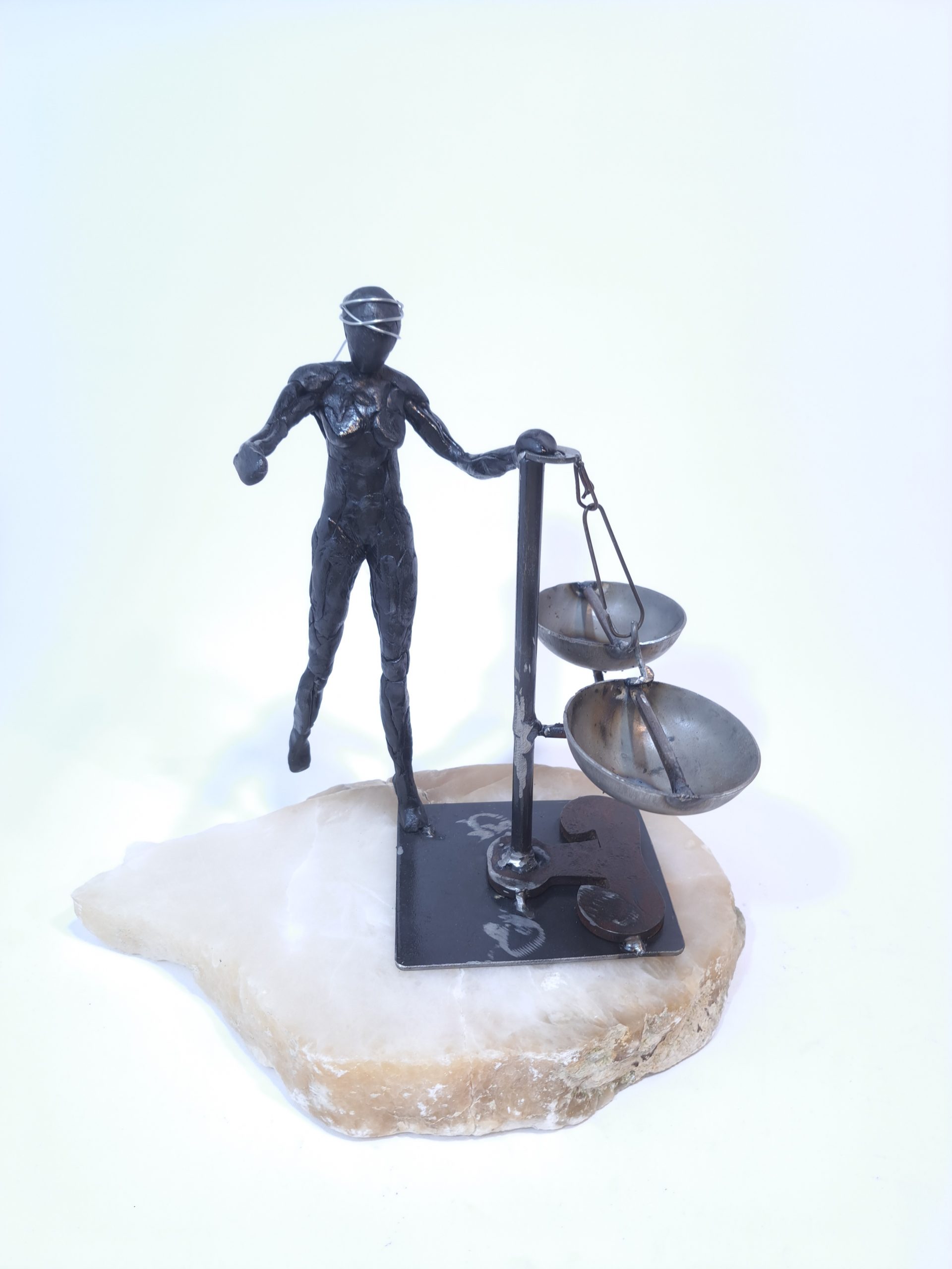 Escultura formada por una figura femenina de resina epoxi con los ojos vendados que sujeta una balanza en equilibro sobre una base de alabastro blanco.