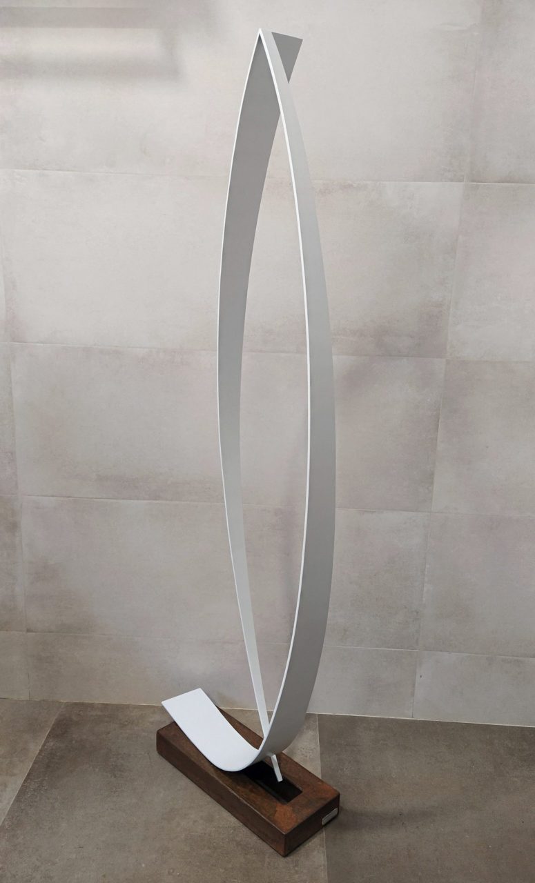 Escultura abstracta de metal con dos piezas curvadas blancas, formando un óvalo en posición vertical, sobre una base rectangular de metal oxidado y barnizado.